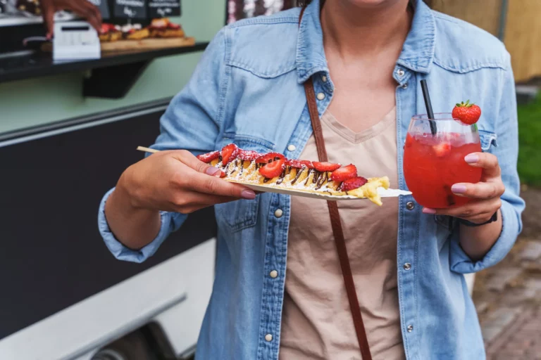 Woman in a denim jacket holding a chocolate and strawberry crepe and a red strawberry iced drink at a food truck window.