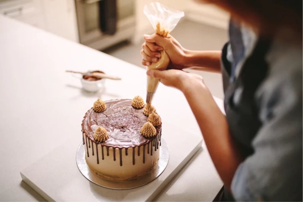 A baker wearing a blue shirt and a black apron pipes decorating frosting on a cake with dripping brown icing while working at a white table in a kitchen.