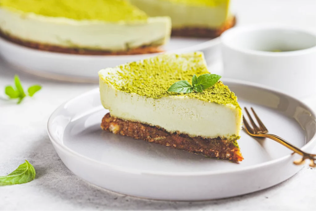 A green matcha cheesecake slice on a white plate with a gold-tone fork, mint leaf garnishes, and a cup of tea, and the full cheesecake in the background.