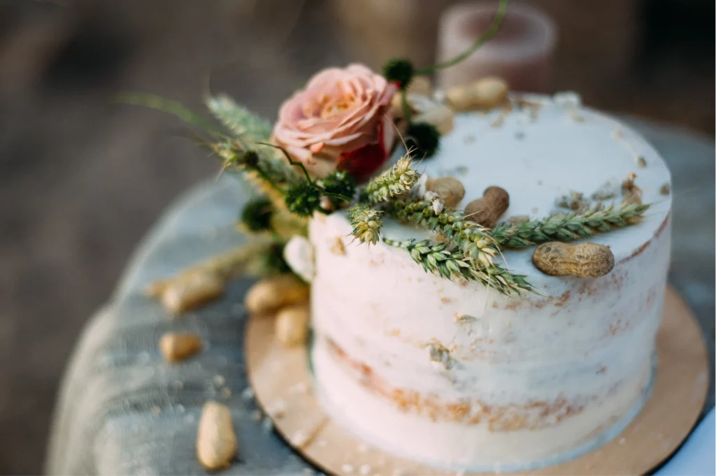 A close-up image of a wedding cake decorated with white frosting, pink flowers, greenery, and peanuts on a table.