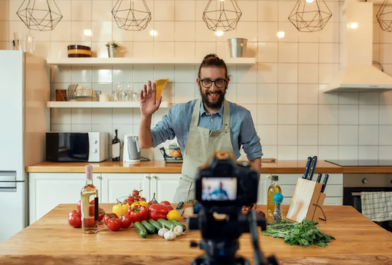 A chef wearing a blue denim shirt and a light gray apron smiles and waves at the camera as he records himself in a modern kitchen. In front of him is an array of vegetables, knives, and condiments set atop a wooden kitchen island.