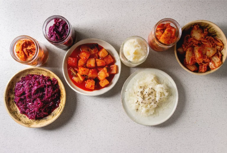 An overhead image of various pickled cabbages and radishes in glass jars and bowls set atop a speckled gray countertop.
