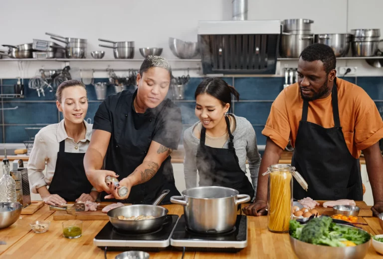 A group of workshop attendees watches a kitchen instructor grind pepper into a skillet during a cooking class.