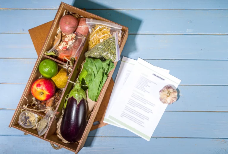 An overhead image of a wooden food subscription box with various vegetables, fruits, and dry goods packed inside. The box is set on a blue slatted floor with instruction cards fanned next to it.