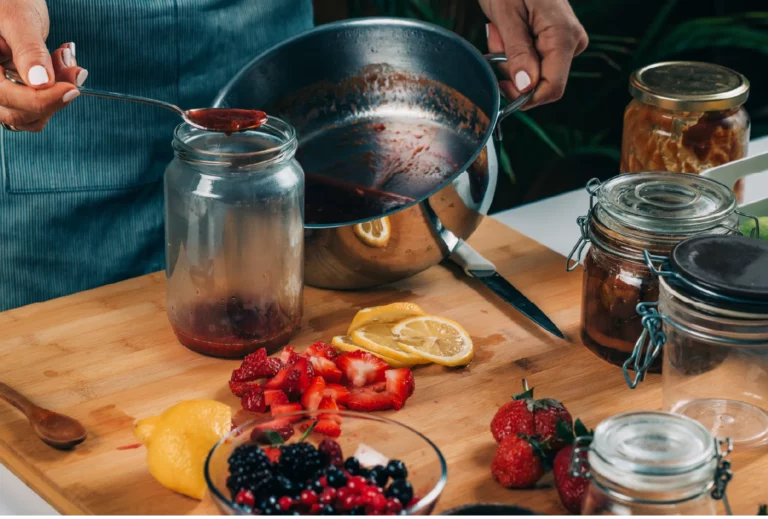 A close-up image of a chef spooning fruit preserves from a steel pot into a glass jar set atop a wooden cutting board with various fruits and jars placed around the working area.