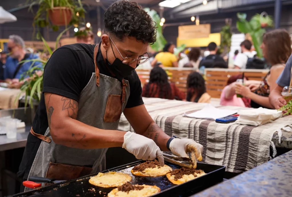 A food vendor wearing a black shirt, gray apron, black face mask, and disposable gloves cooks meat and cheese tostadas on a hot plate in a busy marketplace.