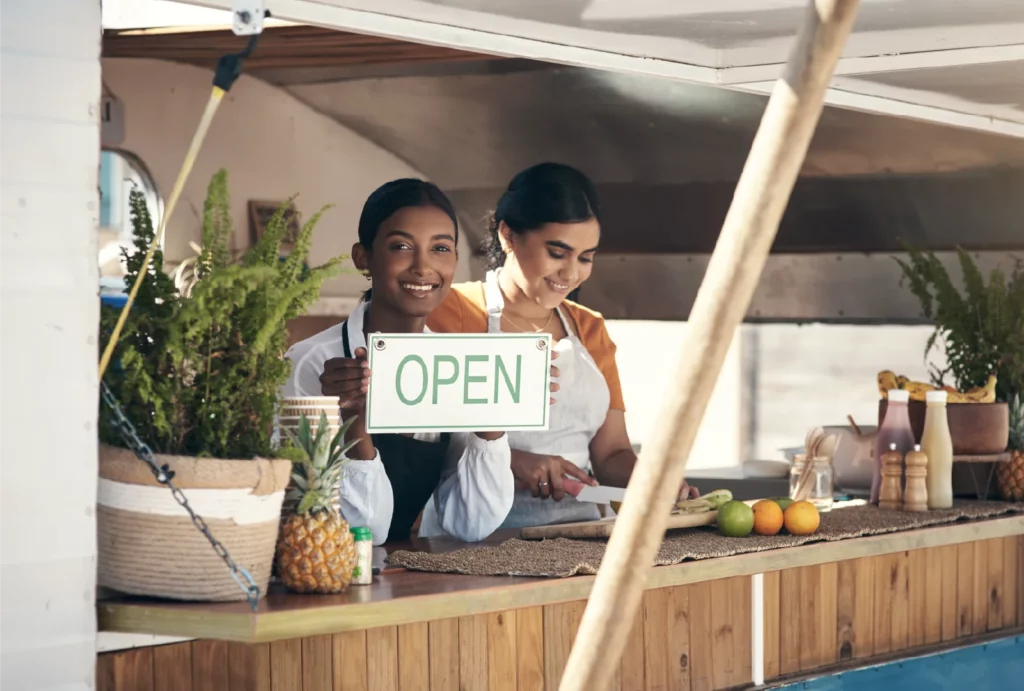 A food vendor wearing a white shirt and black apron holds a green-and-white "open" sign next to her worker, wearing an orange top and a white apron, as they stand in the window of their food truck decorated with green plants, fresh fruit, and rattan placemats.