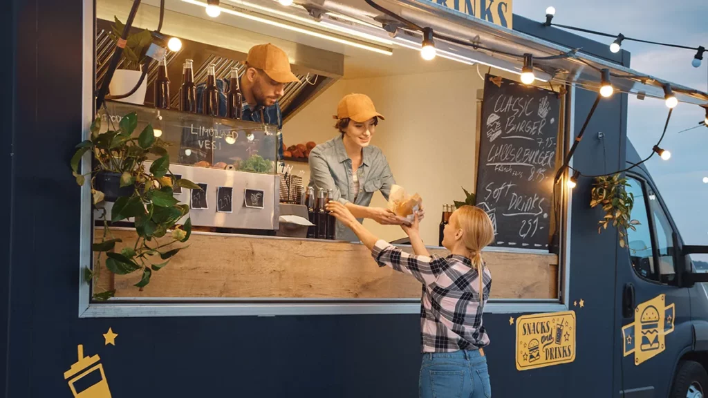 Two employees in yellow hats serve food to a customer out of a black food truck selling snacks and soft drinks.