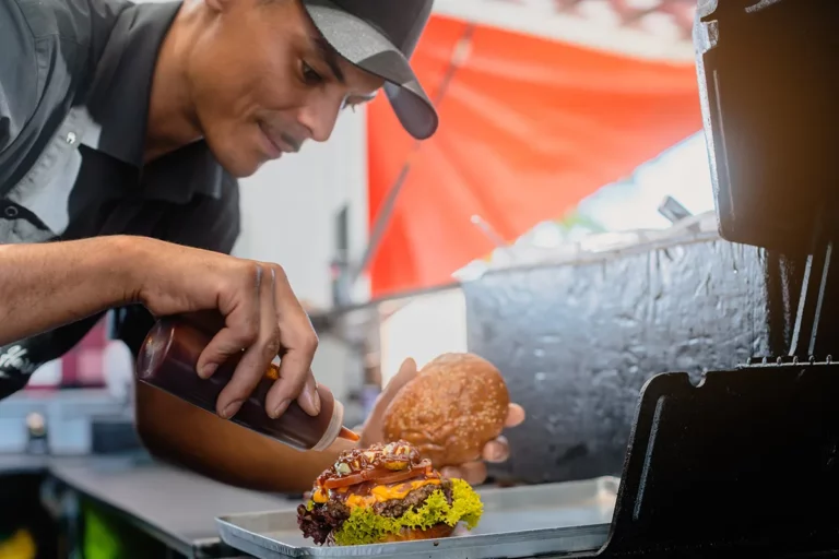 A cook in a food truck adds condiments to a burger before serving it.