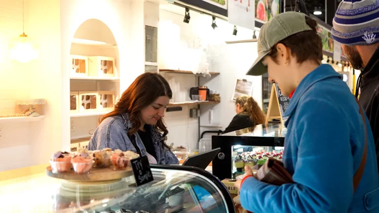 Lauren from Moore Desserts by Julie rings up a customer at her booth with a plate of free samples set up on one of the glass display cases.