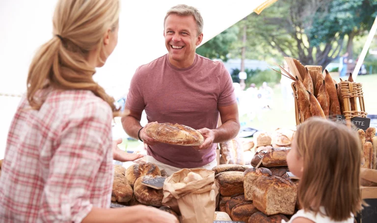 A local baker smiles as he sells a loaf of bread to a family from his bakery stall at a farmers market.