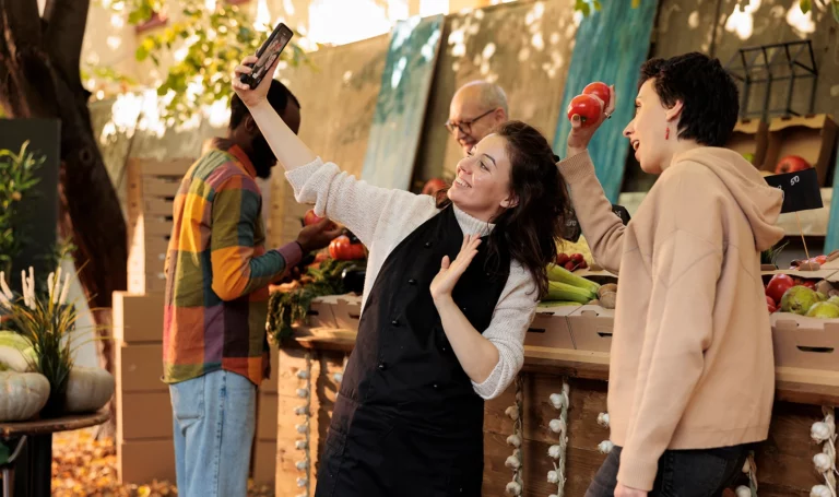 Two shoppers smile and pose with their smart phone as they take a selfie in front of a farmers market booth.