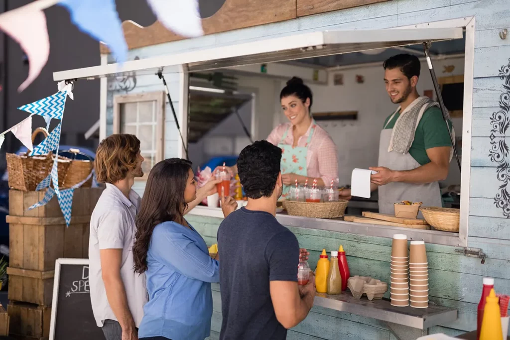 Three adults buying food at a rustic blue food truck with two smiling employees.