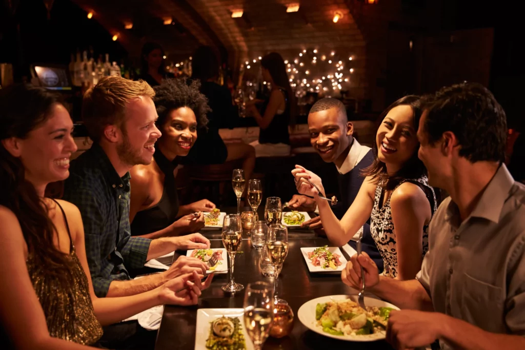 A group of friends laughing, eating, and drinking white wine around a dimly lit restaurant table.
