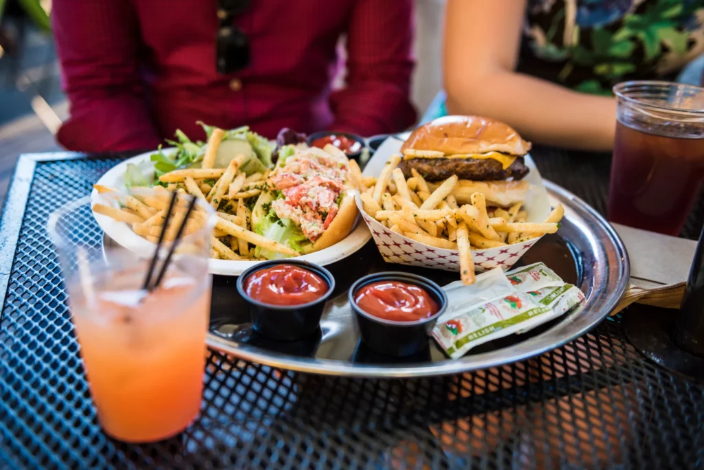 A close-up of American food on a dish at a restaurant's outdoor table, including a lobster roll and a cheeseburger with fries.
