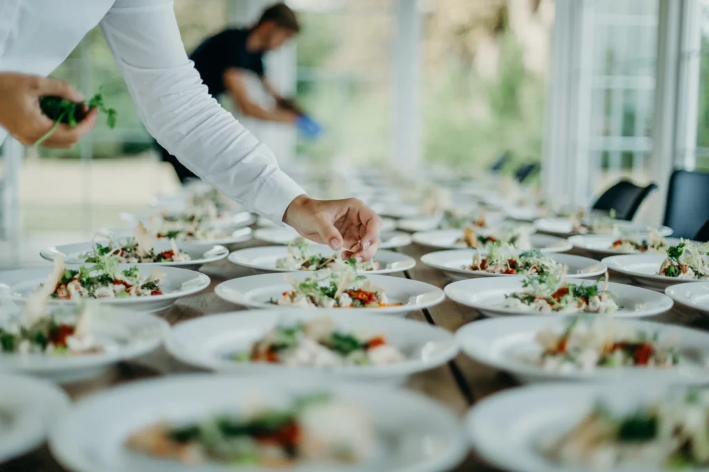 A catering server garnishes a dish set in the middle of multiple identical dishes on a table.