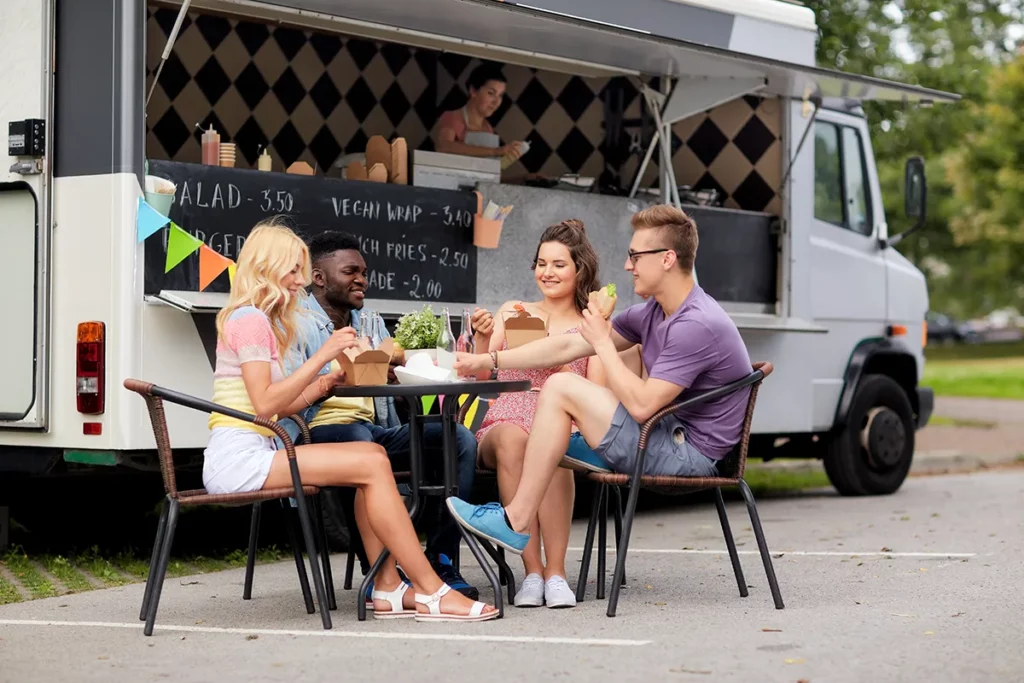 A group of four friends eats at a table outside a food truck parked along the grass at the side of a road.