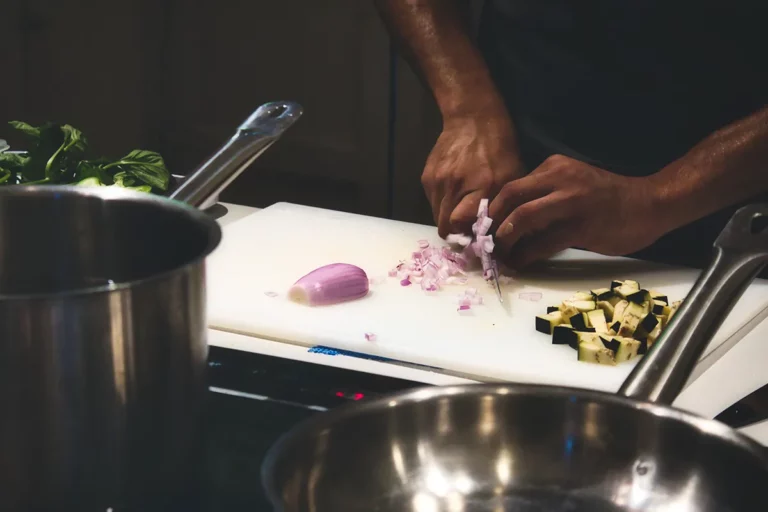 Close-up of a cook dicing a shallot on a cutting board in front of two saucepans.
