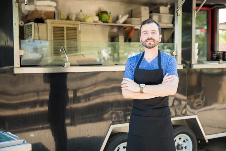 A man in a blue shirt and black apron stands with his arms folded across his chest in front of a black food truck.