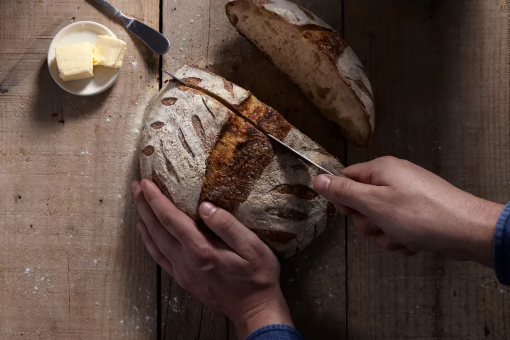 A close-up of a person cutting into a loaf of sourdough bread on a wooden cutting board.