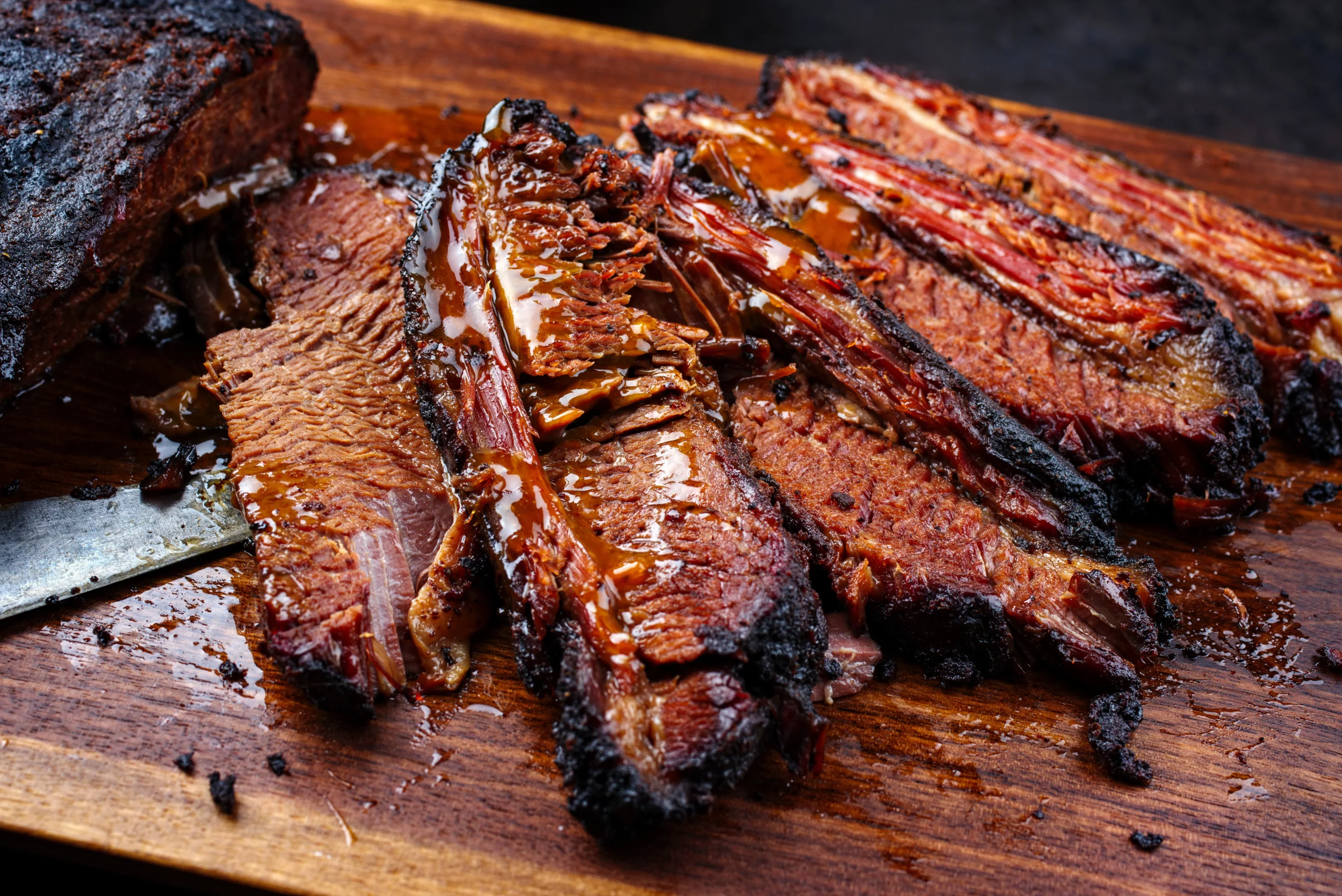 Close-up of sliced smoked brisket with a knife underneath, served on a wooden cutting board.