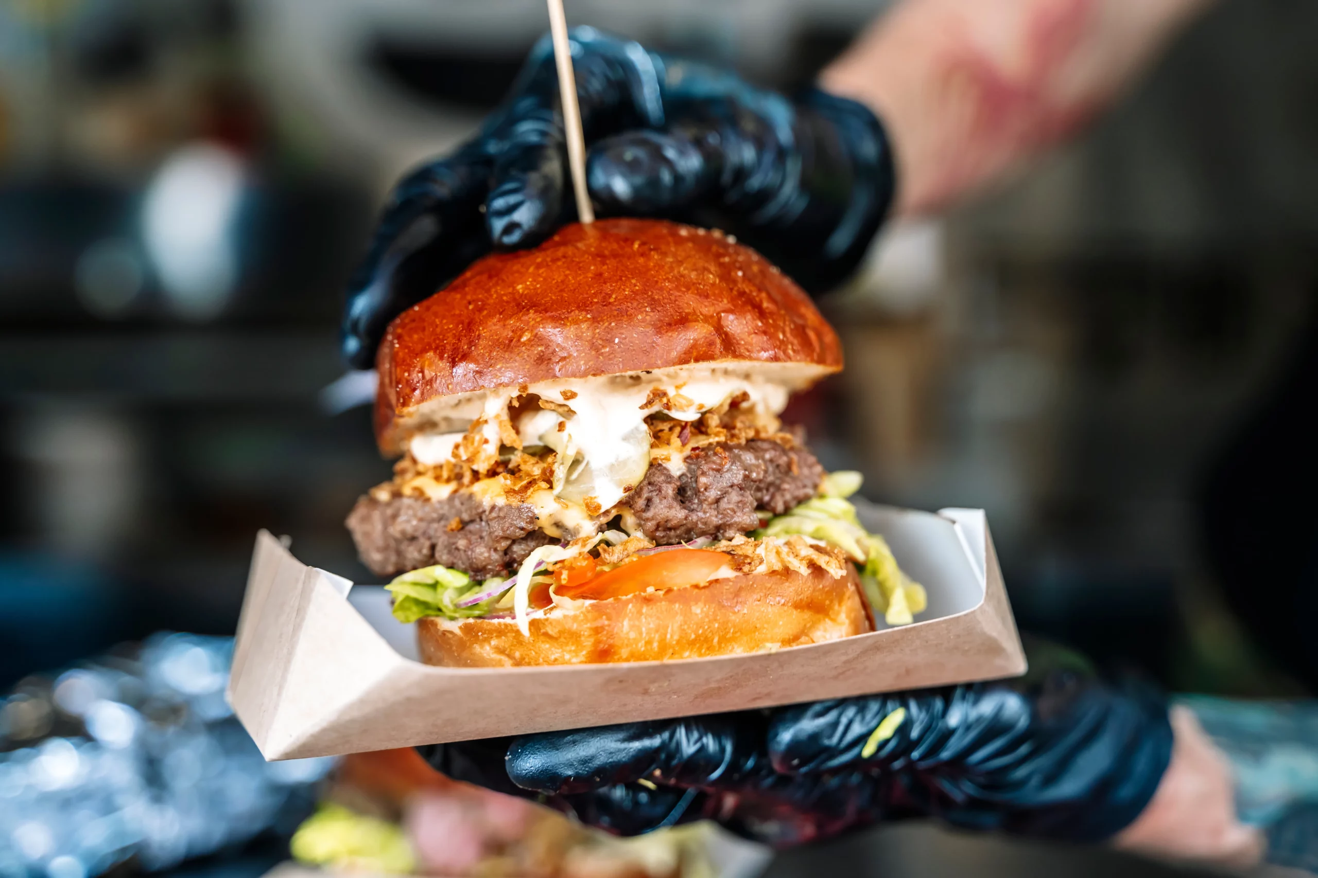 Gloved hand holding an overstuffed cheeseburger with melted cheese and toppings in a food truck serving tray.