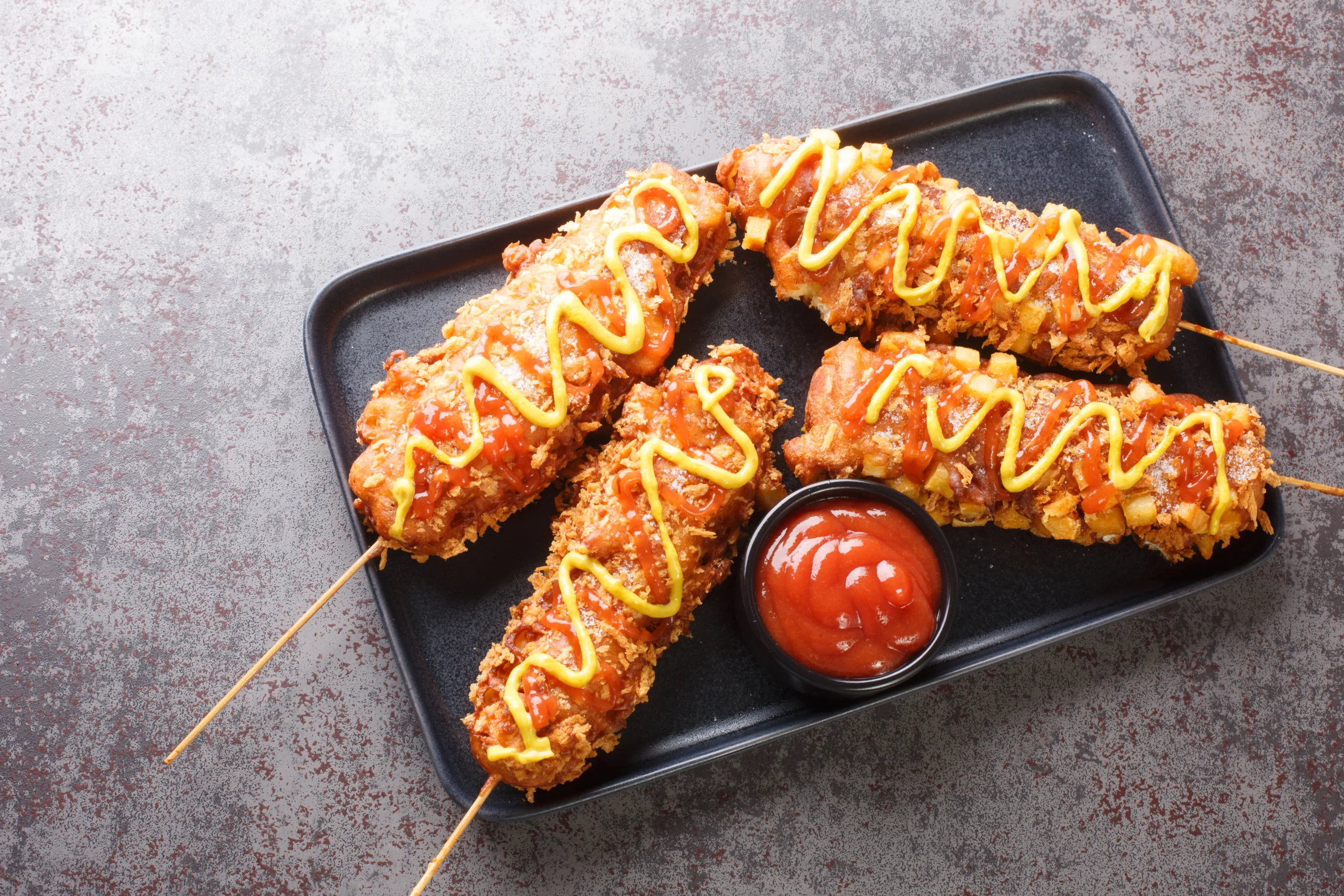 Corn dogs on wooden skewers drizzled with mustard, served with a side of ketchup on a black platter.