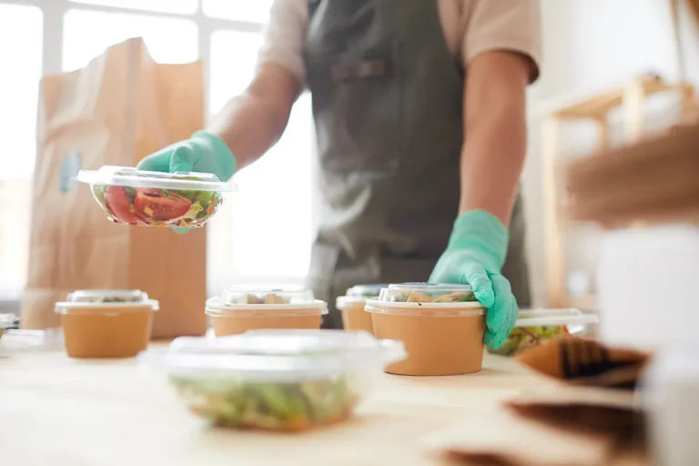 A food service worker wears a black apron and food-safe gloves while handling packaged salads for delivery.