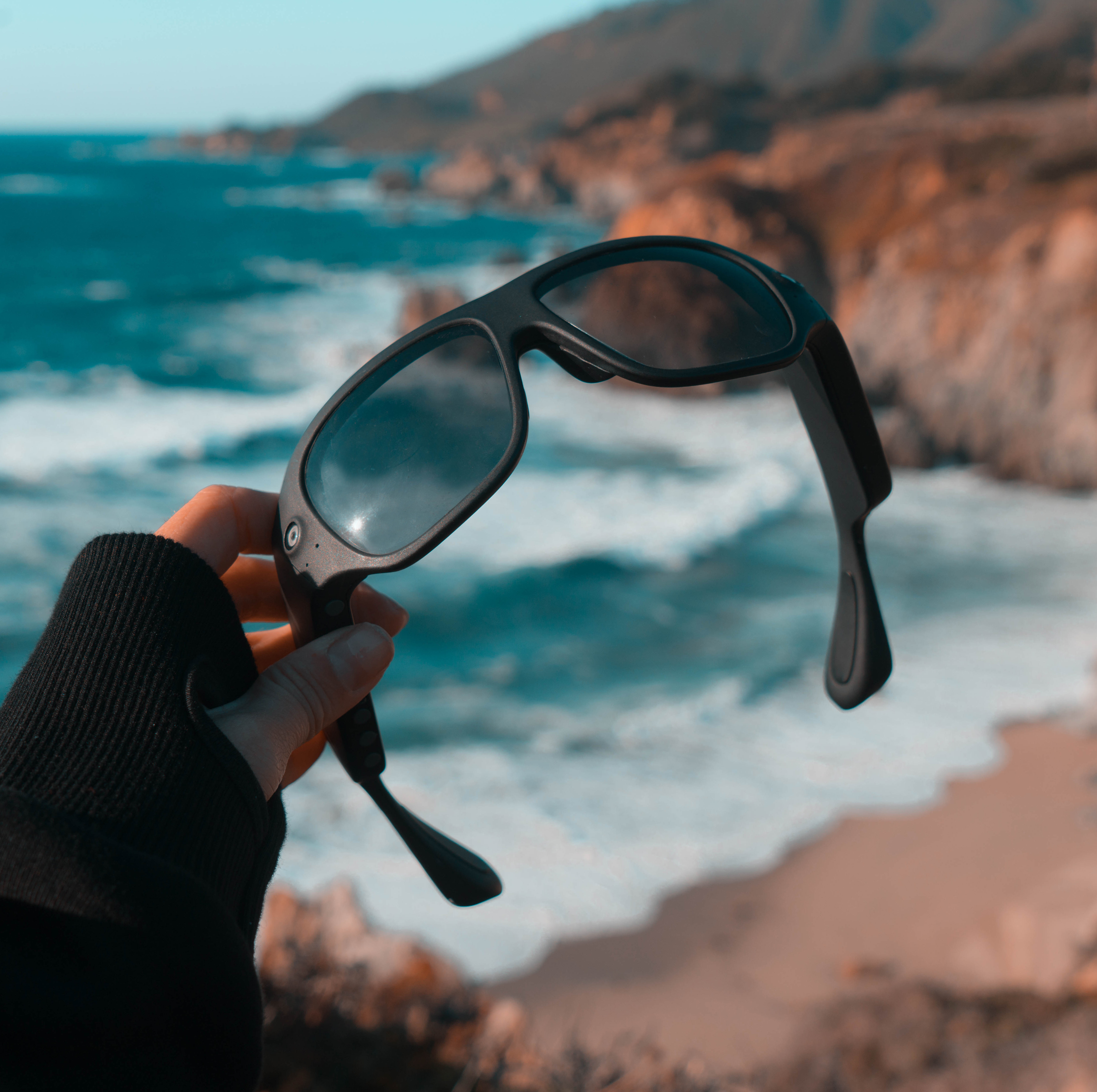 hand holding a pair of orbi sunglasses with beach surf in background