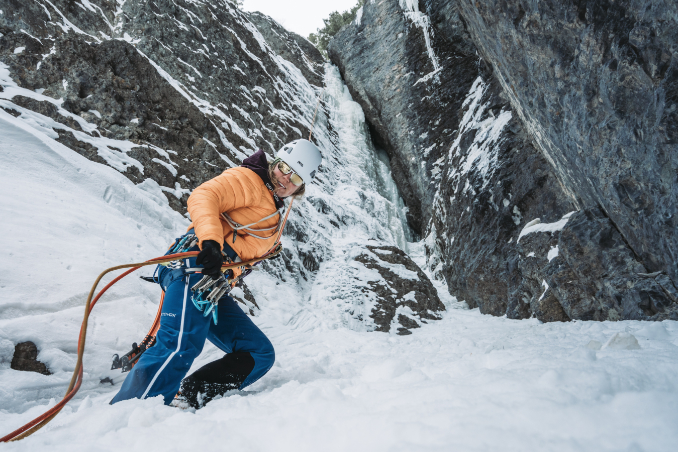 a roped up female climber wearing a pale coral puffy jacket and white climbing helmet as she ascends a gully in winter