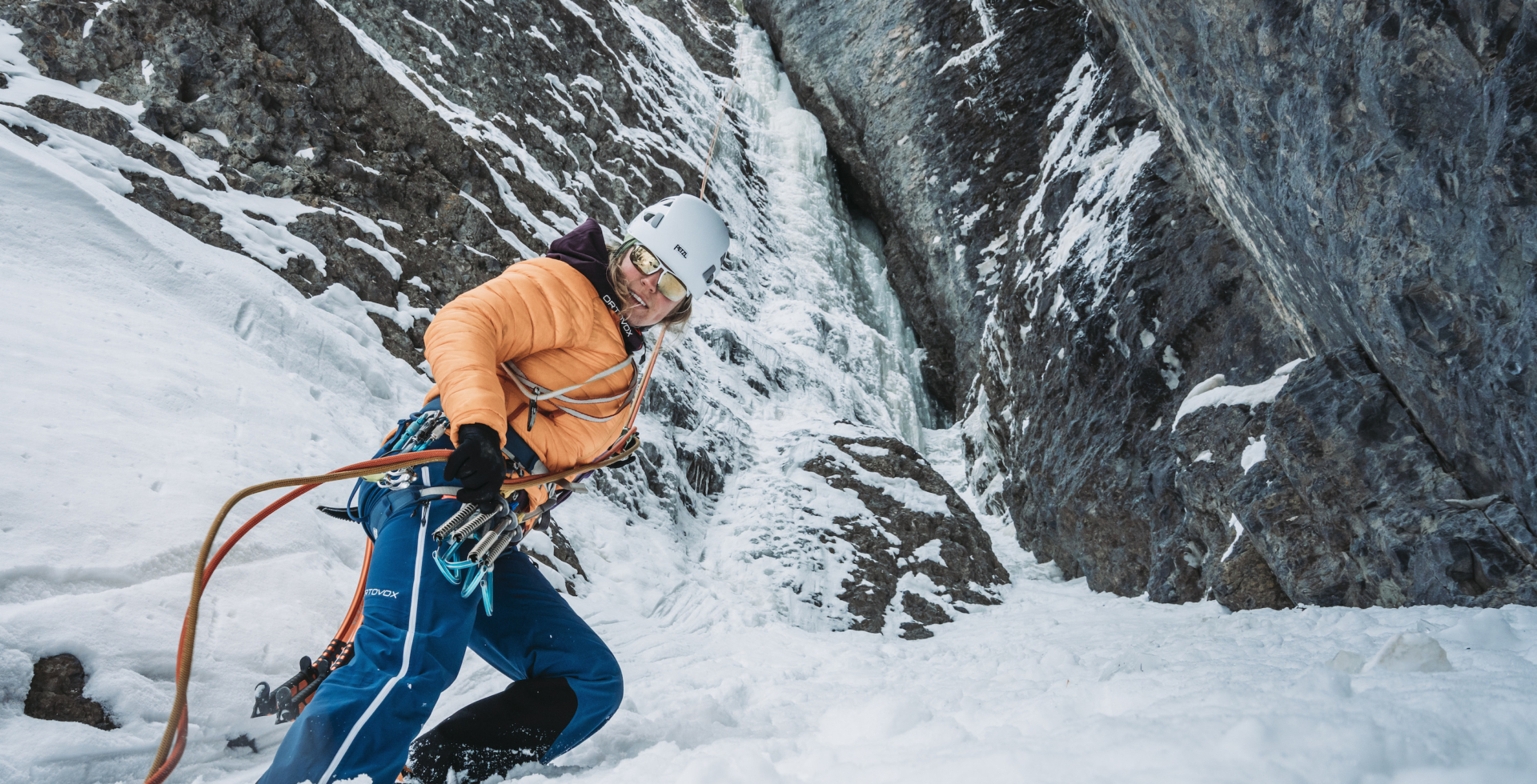 a roped up female climber wearing a pale coral puffy jacket and white climbing helmet as she ascends a gully in winter