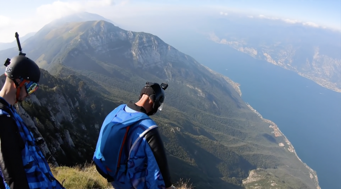two men preparing to wingsuit off very tall mountain with view of water below