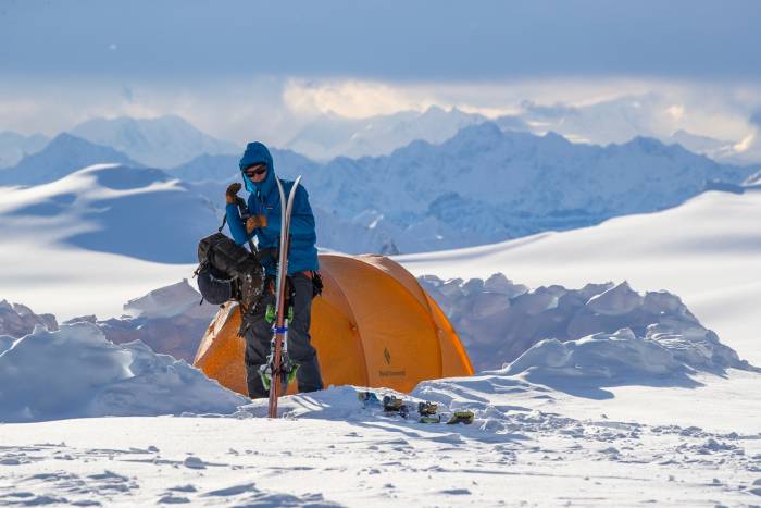 Man on snowy landscape carrying backpack and gear towards pair of skis with orange tent in background