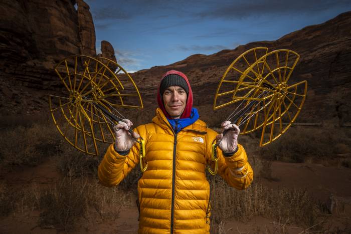 Alex Honnold in a yellow puffy and beanie holding giant 2-foot cams