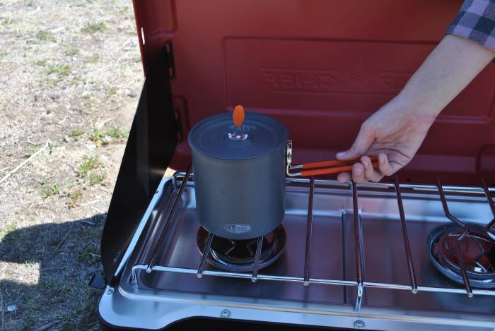 Mary wearing a flannel sleeve, reaching for a pot handle cooking over a camp chef stove