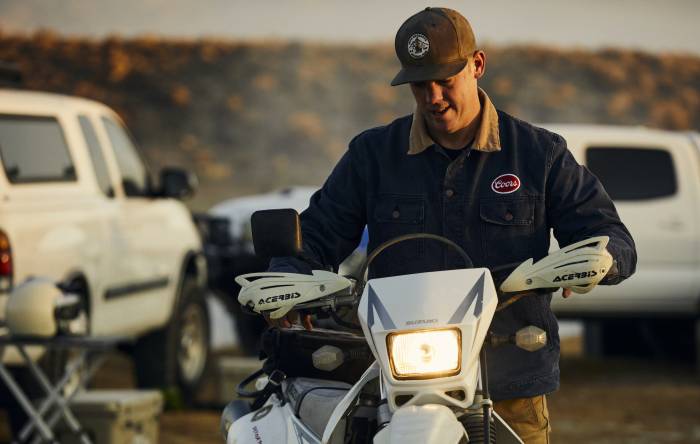 Man wearing Coors x Huckberry series hat and jacket mounting a white motorbike