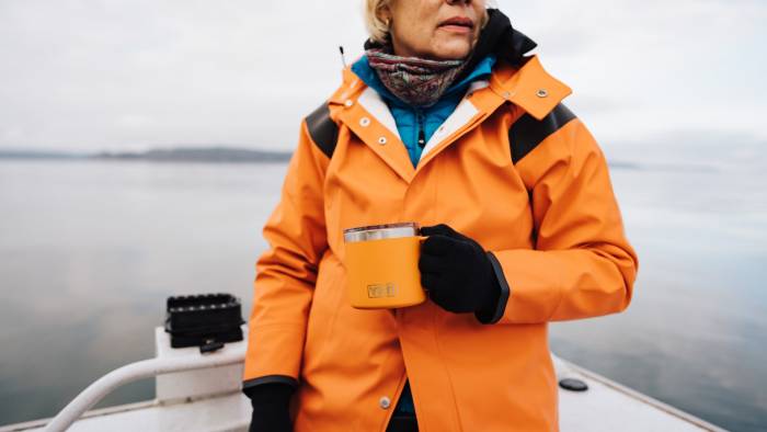 older female fisherman on a boat wearing orange shell jacket and holding king crab YETI mug