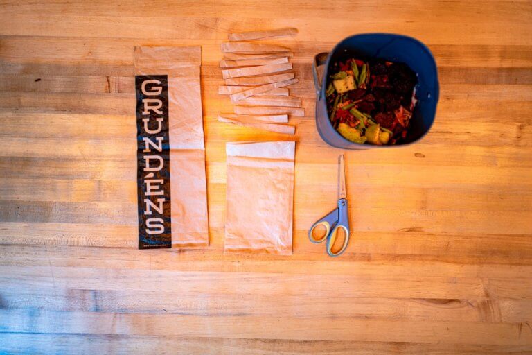 Grundéns Ditches Poly Bags, Launches Compostable Packaging for Sustainability Compostable poly bags with the Grundéns’ logo next to a home compost bin on a wooden table