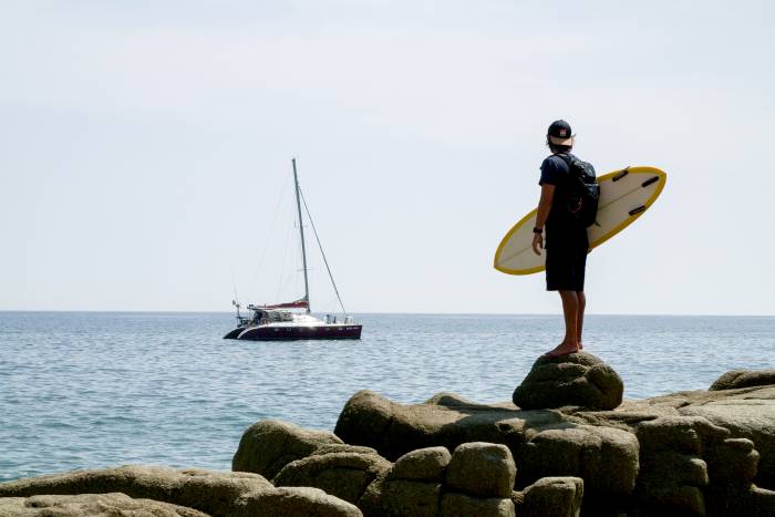 a surfer standing on a rocky shore looking out to a sailboat