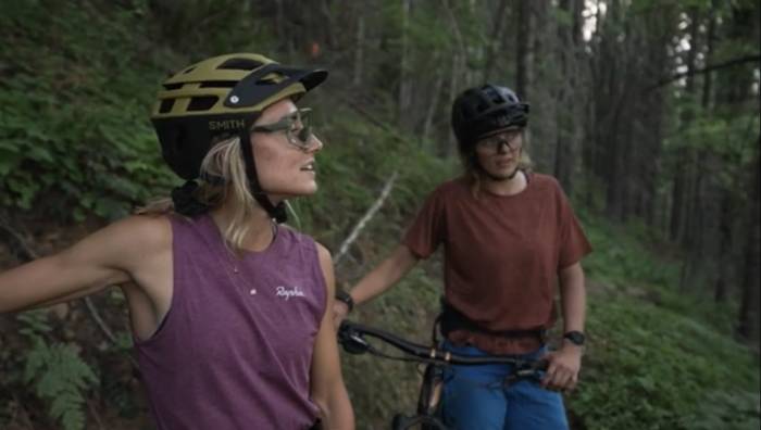 two women in short sleeves and bike helmets looking off at a trail in the distance