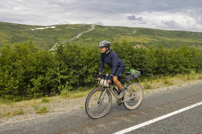 Lael Wilcox gravel riding in Alaska alone a road with a oil pipeline up a mountain in the background
