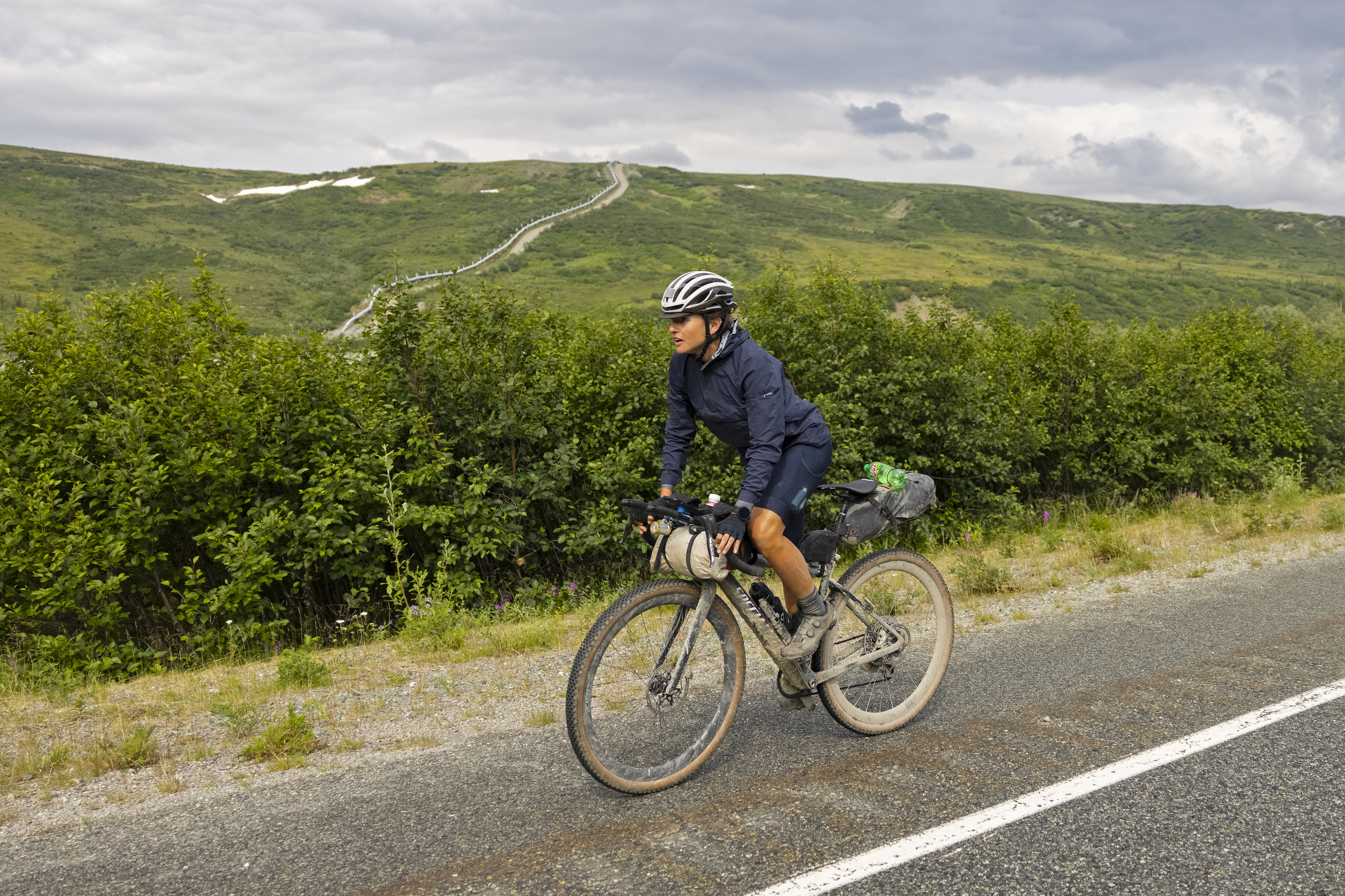 Lael Wilcox gravel riding in Alaska alone a road with a oil pipeline up a mountain in the background