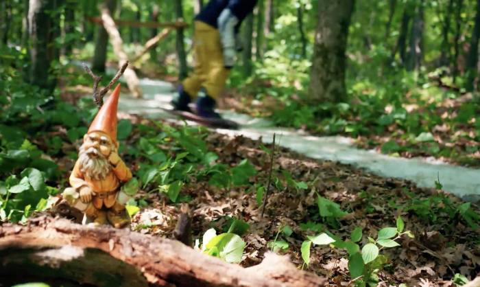 gnome in foreground with snowboarder cruising through green forest in background