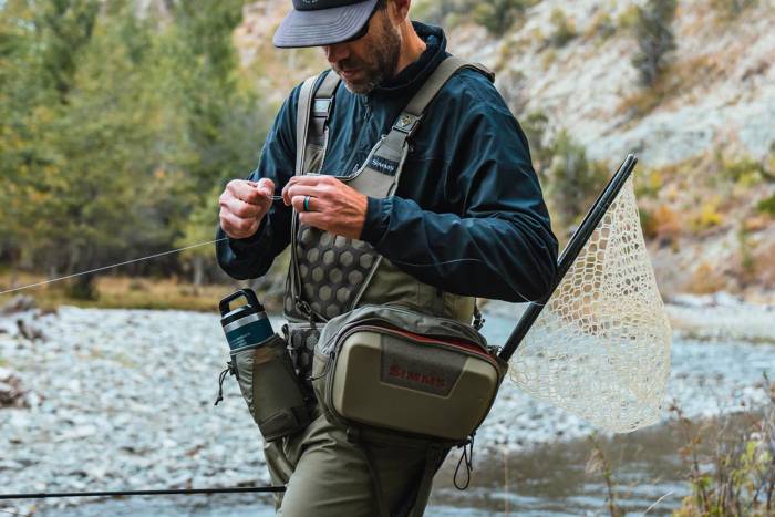 Fly fisherman standing in a stream tying a knot wearing the SIMMS Fishing Flyweight Hybrid Hip System and Flyweight Waders