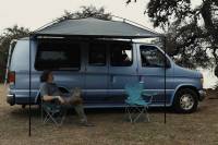 man with shoulder-length hair resting in a chair under a Moon Fab shade attached to his campervan