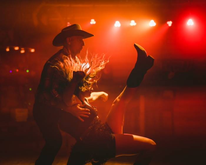 man and woman in cowboy hats and boots dancing under a red-lit stage