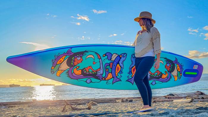 Women with a sun hat on and jeans carrying a Carta stand up paddle board by Sea Gods.