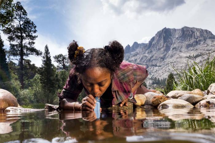 Women with hair up and wearing a flannel drinking water in a stream with a LifeStraw.