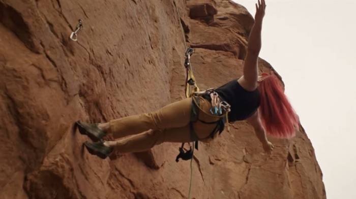 a female climber with bright pink hair hanging off a rope with her arms outstretched