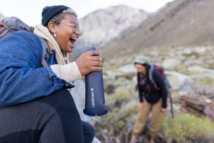 woman laughing while holding her LifeStraw Squeeze 1L with a friend in the background in the mountains.
